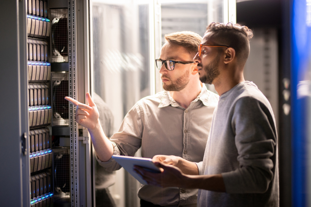 Two IT support technicians holding a tablet and looking at a server mainframe trying to diagnose an issue